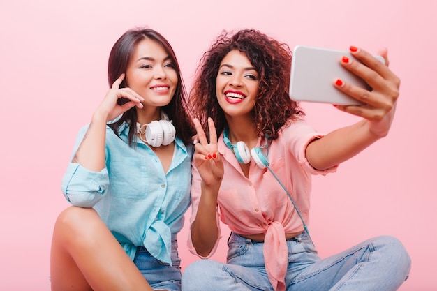 Feliz niña africana con cara bonita posando con el signo de la paz junto a una encantadora amiga. aforable mujer mulata en jeans y camisa rosa haciendo selfie con elegante dama hispana.