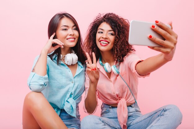 Feliz niña africana con cara bonita posando con el signo de la paz junto a una encantadora amiga. Aforable mujer mulata en jeans y camisa rosa haciendo selfie con elegante dama hispana.