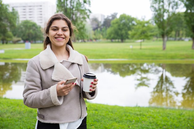 Feliz niña alegre con teléfono disfrutando de un café para llevar