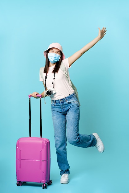 Feliz niña asiática en máscara médica turista posando emocionado con maleta que va de vacaciones viajando un ...