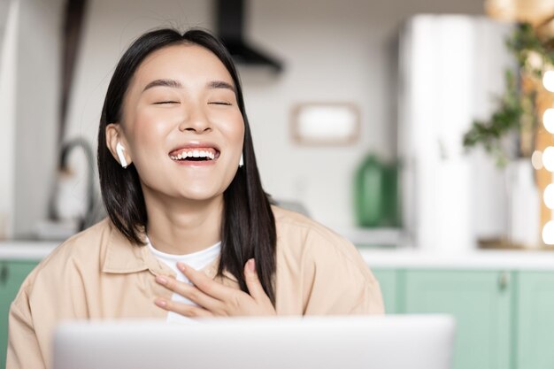 Feliz niña asiática riendo viendo un video o teniendo una llamada a través de una aplicación portátil que trabaja de forma remota ...