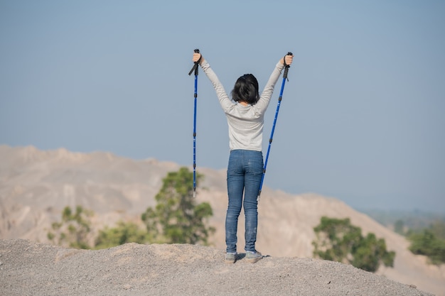 Feliz niña asiática senderismo en las montañas de pie sobre una cresta rocosa cumbre y un poste con vistas al paisaje.