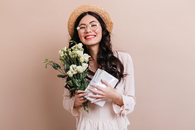 Feliz niña asiática sosteniendo presente y flores Sonriente mujer china con sombrero posando con ramo de eustoma