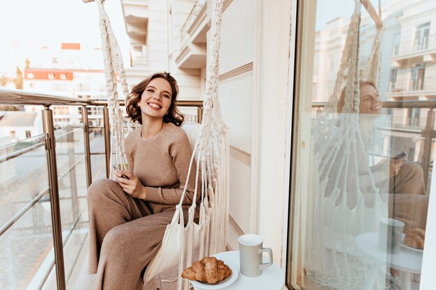 Feliz niña blanca sentada en la terraza con croissant sabroso. Foto de mujer joven riendo desayunando en el balcón.