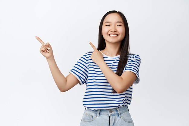 Feliz niña bonita asiática señalando con el dedo a la izquierda y sonriendo en blanco.