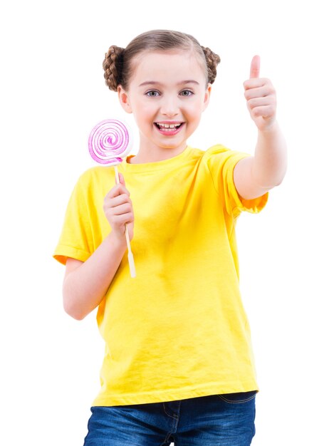Feliz niña bonita en camiseta amarilla con caramelos de colores que muestran los pulgares para arriba signo - aislado en blanco.