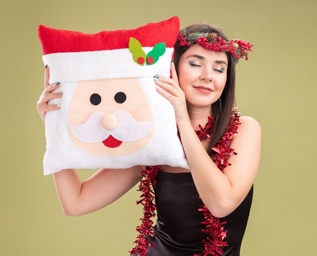 Feliz niña bonita caucásica con corona de Navidad y guirnalda de oropel alrededor del cuello sosteniendo la almohada de santa claus tocando la cabeza con los ojos cerrados aislado sobre fondo verde oliva