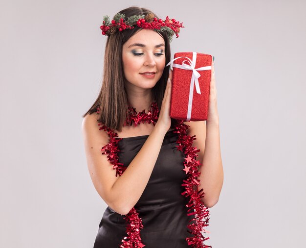 Feliz niña bonita caucásica con corona de Navidad y guirnalda de oropel alrededor del cuello sosteniendo y mirando el paquete de regalo aislado sobre fondo blanco con espacio de copia