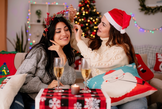 Feliz niña bonita con gorro de Papá Noel sostiene y mira a su amiga holly wreath sentada en sillones y disfrutando de la Navidad en casa