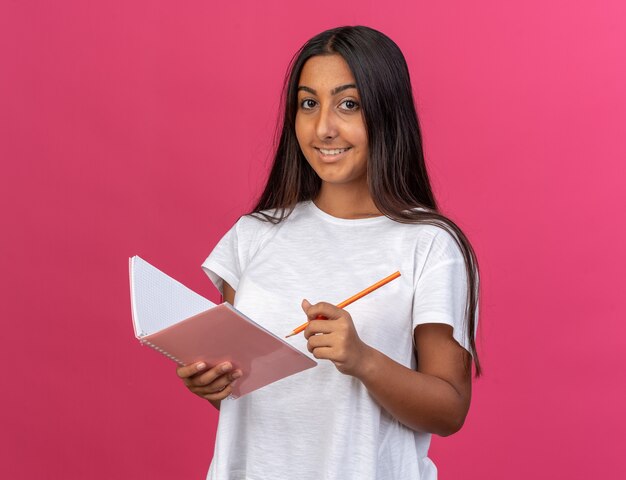 Feliz niña en camiseta blanca con cuaderno y lápiz mirando a la cámara con una sonrisa en la cara de pie sobre rosa