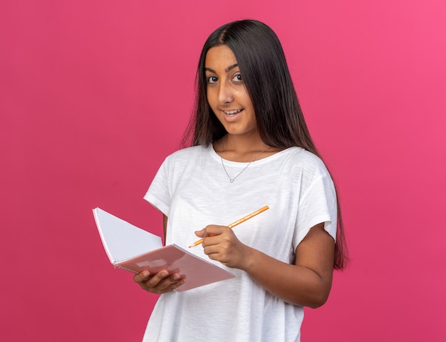 Feliz niña en camiseta blanca con cuaderno y lápiz mirando a la cámara con una sonrisa en la cara