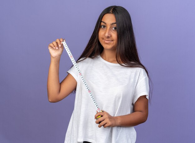 Feliz niña en camiseta blanca sosteniendo cinta métrica mirando a la cámara con una sonrisa en la cara de pie sobre fondo azul.