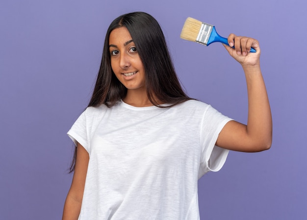 Feliz niña en camiseta blanca sosteniendo un pincel mirando a la cámara sonriendo alegremente de pie sobre azul