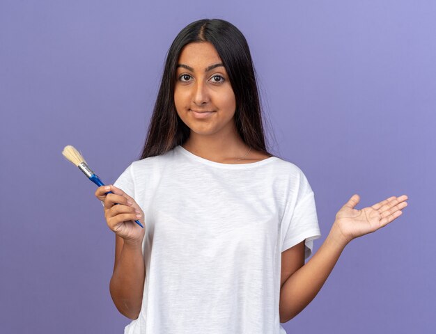 Feliz niña en camiseta blanca sosteniendo un pincel mirando a la cámara con una sonrisa en la cara de pie sobre fondo azul.