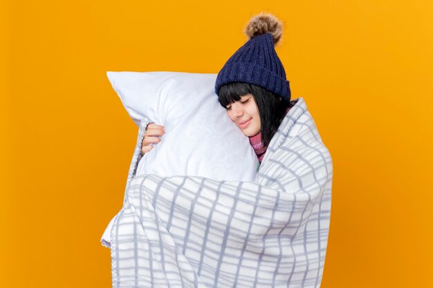 Feliz niña caucásica enferma joven con gorro de invierno y bufanda envuelta en tela escocesa sosteniendo la almohada tocando la cara con los ojos cerrados aislado sobre fondo naranja con espacio de copia