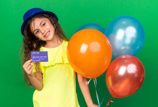 Feliz niña caucásica con gorro de fiesta azul sosteniendo globos de helio y tarjeta de crédito aislado en la pared verde con espacio de copia