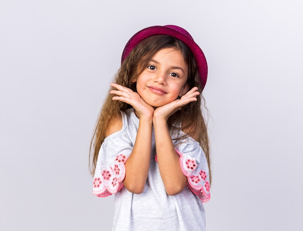 Feliz niña caucásica con gorro de fiesta morado poniendo las manos en el mentón aislado en la pared blanca con espacio de copia