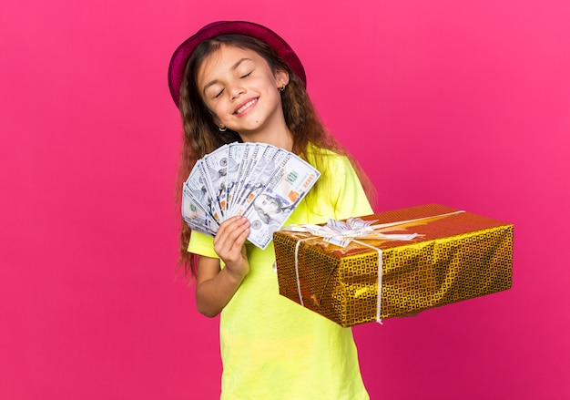 Feliz niña caucásica con gorro de fiesta púrpura con caja de regalo y dinero aislado en la pared rosa con espacio de copia