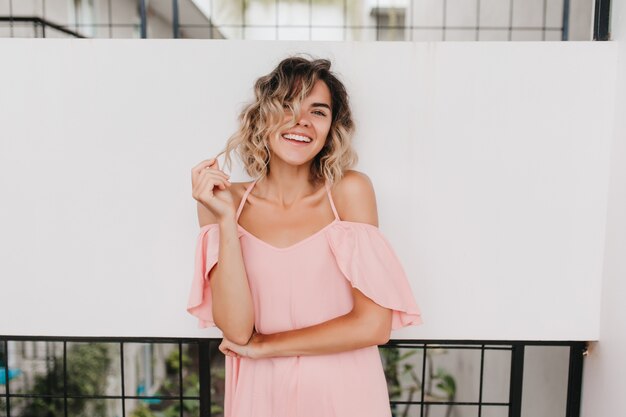 Feliz niña caucásica jugando con su cabello ondulado. Linda mujer de pelo corto con piel bronceada lleva un vestido rosa de verano.