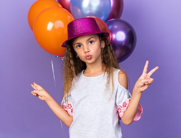 Feliz niña caucásica con sombrero de fiesta violeta gesticulando el signo de la victoria de pie delante de los globos de helio aislados en la pared púrpura con espacio de copia