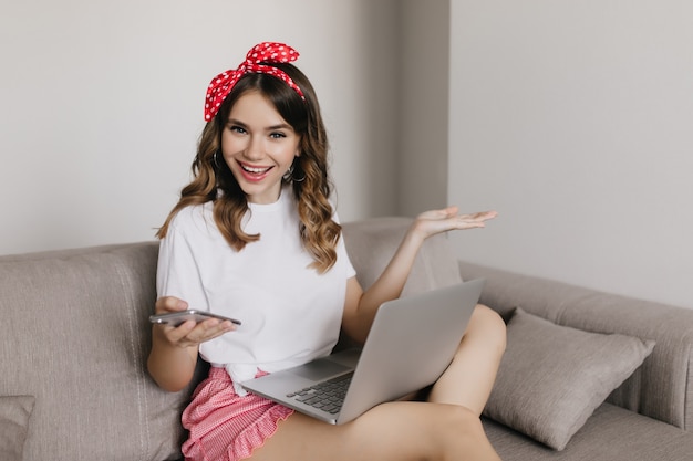 Feliz niña caucásica con teléfono en mano sentado en el sofá en su habitación. Retrato interior de dama sensual con portátil posando en el sofá.