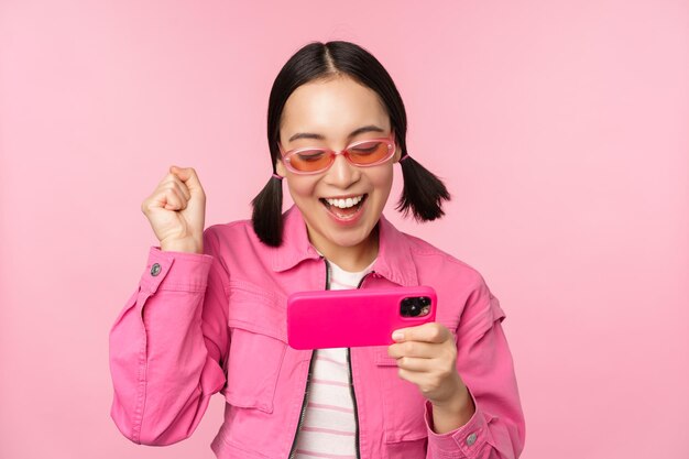 Feliz niña coreana sonriente ganando en el teléfono móvil mirando la pantalla horizontal del teléfono inteligente y regocijándose por lograr el objetivo celebrando de pie sobre un fondo rosa