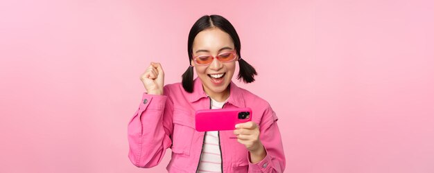 Feliz niña coreana sonriente ganando en el teléfono móvil mirando la pantalla horizontal del teléfono inteligente y regocijándose por lograr el objetivo celebrando de pie sobre un fondo rosa