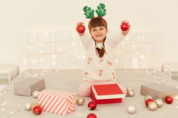 Feliz niña emocionada con suéter blanco y cuernos de ciervo verde de fiesta posando con los brazos levantados con bolas rojas de Navidad, mirando a la cámara con expresión feliz.