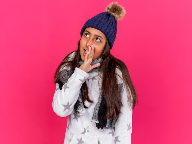 Feliz niña enferma joven mirando al lado con sombrero de invierno con bufanda susurrando aislado sobre fondo de color rosa