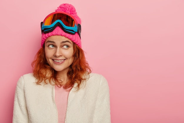 Foto gratuita feliz niña esquiadora sonríe suavemente, enfocada a un lado, usa gorro de invierno rosa con pompón, suéter blanco suave en la cremallera, gafas de esquí en la cabeza, posa contra la pared rosada