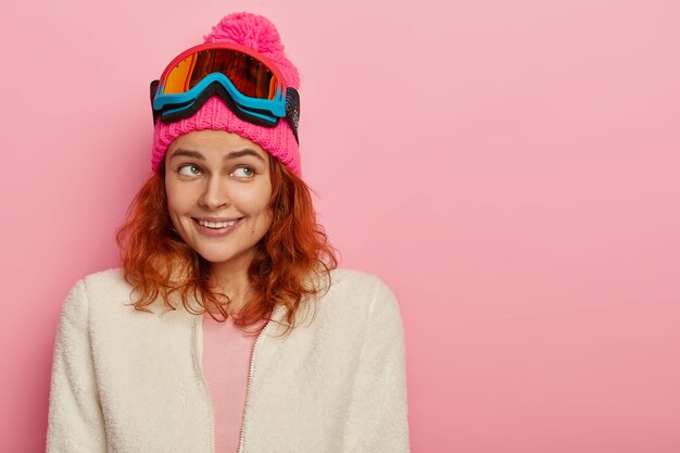 Feliz niña esquiadora sonríe suavemente, enfocada a un lado, usa gorro de invierno rosa con pompón, suéter blanco suave en la cremallera, gafas de esquí en la cabeza, posa contra la pared rosada