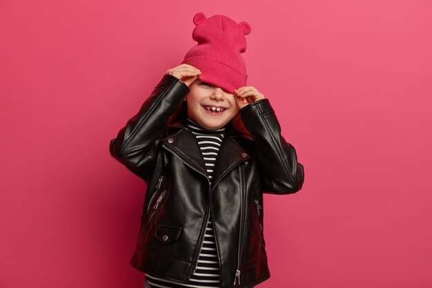 Feliz niña europea usa sombrero rosa con orejas, chaqueta de cuero negro, se esconde cuando la madre intenta hacer una foto de ella, tiene una expresión alegre, posa sobre una pared rosa vibrante. Estilo e infancia