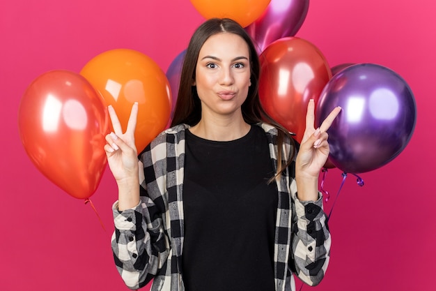 Feliz niña hermosa de pie delante de globos mostrando gesto de paz aislado en la pared rosa