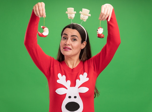 Feliz niña hermosa vistiendo un suéter de Navidad con juguetes de levantamiento de aro de pelo de Navidad aislado sobre fondo verde