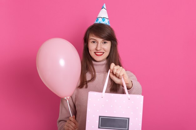 Feliz niña morena con globo rosa y cumpleaños presente en las manos, mantiene la boca abierta, sorprendido