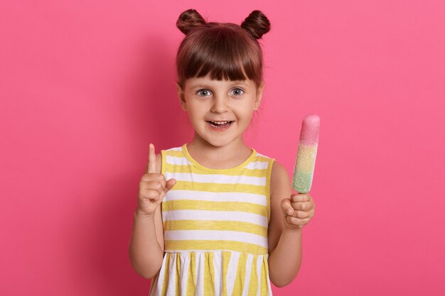 Feliz niña niño sonriente con helado y apuntando hacia arriba con su dedo índice, vestido de verano con rayas blancas y amarillas.