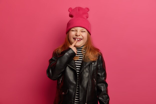 Feliz niña pelirroja con ropa fashionabe, indica su nuevo diente, tiene una infancia inolvidable, entrecierra los ojos con placer, posa contra una pared rosa brillante. Concepto de crecimiento de niños