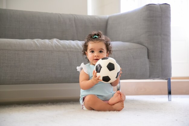 Feliz niña de pelo rizado negro con ropa azul pálido sentada en el suelo en casa, mirando a otro lado, jugando al fútbol. Vista frontal. Niño en casa y concepto de infancia.