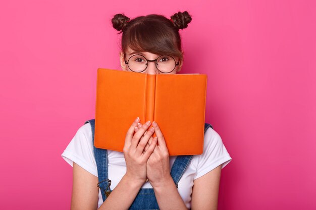 Feliz niña pensativa de pie aislado en rosa, sosteniendo el cuaderno naranja, cubriendo la mitad de la cara, mirando atentamente a la cámara, teniendo racimos, vistiendo ropa casual de moda.