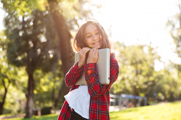 Feliz niña de pie en el parque mientras abraza a su computadora portátil