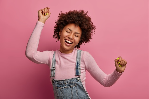 Feliz niña de piel oscura disfruta de cada momento de la vida, baila y se mueve, levanta los brazos y aprieta los puños, cierra los ojos, tiene buen humor, usa denim sarafan y cuello alto, aislado en la pared rosa