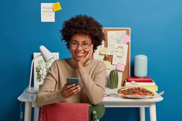 Feliz niña de piel oscura disfruta de Internet de alta velocidad gratis, usa el teléfono móvil para enviar mensajes de texto, se sienta en el lugar de trabajo