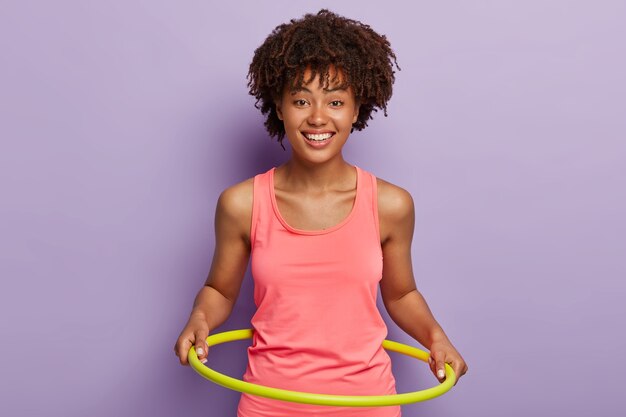 Feliz niña de piel oscura gira hula hoop, disfruta de ejercicios activos y entrenamiento gimnástico, usa equipo de entrenamiento