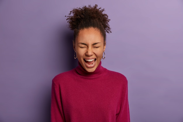 Foto gratuita feliz niña de piel oscura no puede contener la risa, entrecierra los ojos, se ríe por una broma divertida, mira algo adorable o gracioso, usa un cuello alto de color burdeos, aislado en la pared púrpura, tiene un día positivo