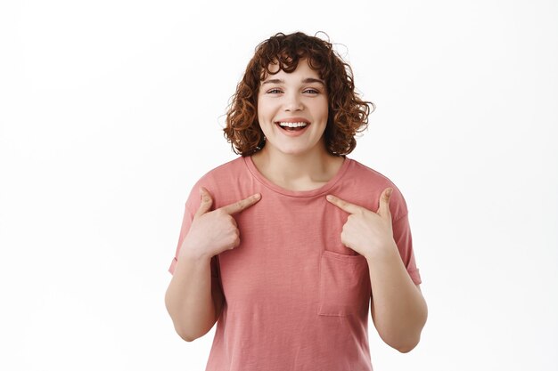 Feliz niña riendo apuntando a sí misma y sonriendo de pie en camiseta en blanco