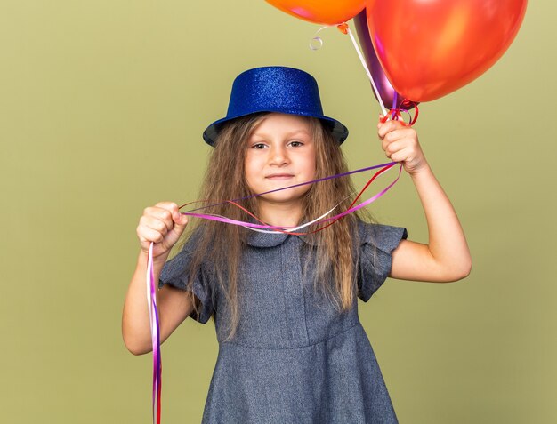 Feliz niña rubia con gorro de fiesta azul sosteniendo globos de helio aislado en la pared verde oliva con espacio de copia