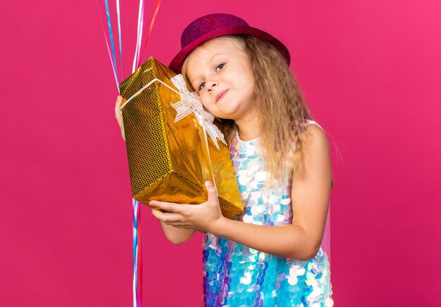 Feliz niña rubia con gorro de fiesta púrpura sosteniendo globos de helio y caja de regalo aislada en la pared rosa con espacio de copia