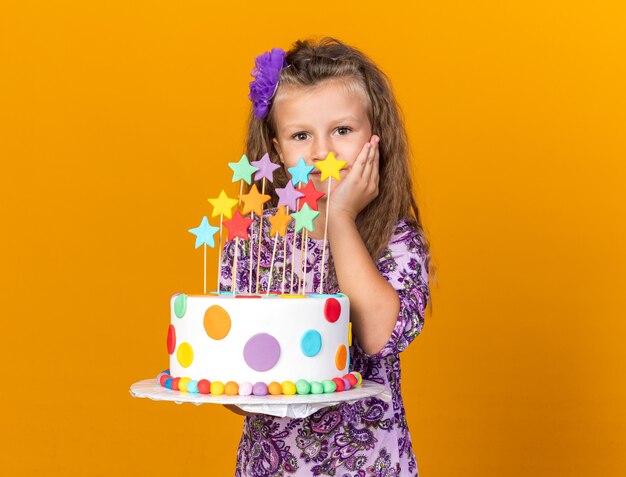 Feliz niña rubia sosteniendo la torta de cumpleaños y poniendo la mano en la cara aislada en la pared naranja con espacio de copia