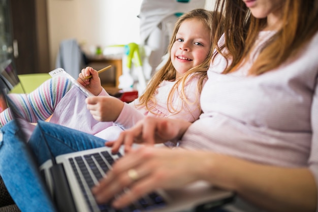 Feliz niña sentada al lado de su madre usando la computadora portátil