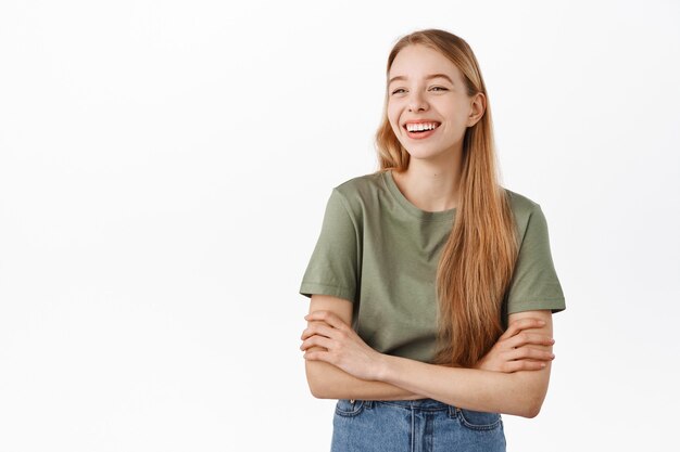 Feliz niña sincera riendo y sonriendo, mirando a un lado el logotipo del anuncio con cara de satisfacción, de pie como si tuviera una conversación informal y relajante con amigos, pared blanca